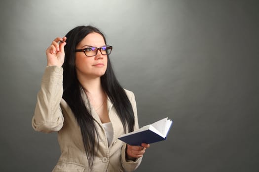 the thoughtful girl, the brunette wearing spectacles with the handle and a notebook on a gray background, looks up