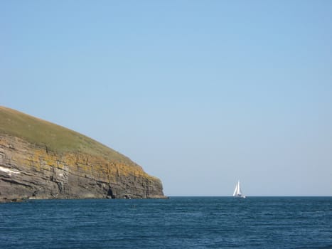 A yacht sailing into the distance around a headland cliff with blue sea and sky.