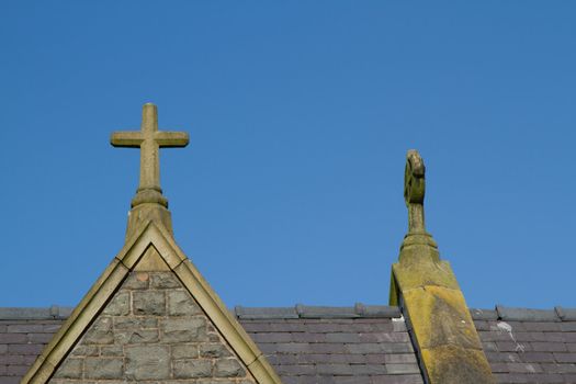 A stone carved chistian cross on an apex of a roof with a blue sky in the background.