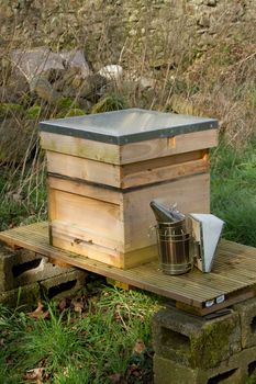 A wooden bee hive on a stand in a shaded space with a metal smoker.