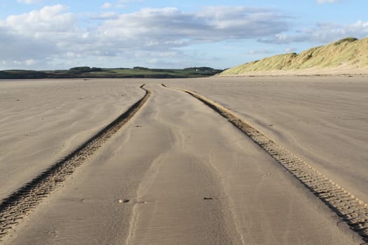 Tire tracks going into the distance on a beach with a sand dune and green fields in the distance backed by a blue cloudy sky.