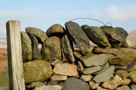 A dry stone wall construction with layers of rock and a coping with rusty wire against a wooden post.