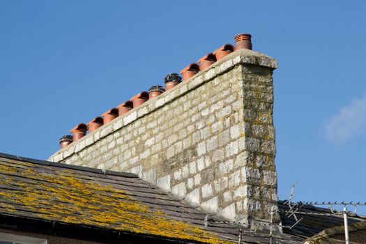 An array of chimney pots on a stone built stack on a slate roof with yellow lichen against a blue sky.