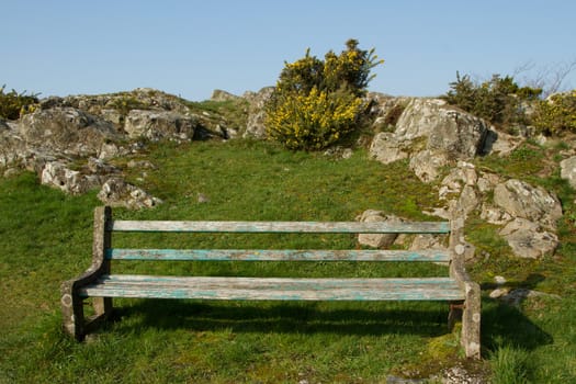 An old bench with flaked blue paint on a patch of green grass with rocks and gorse plants against a blue sky.