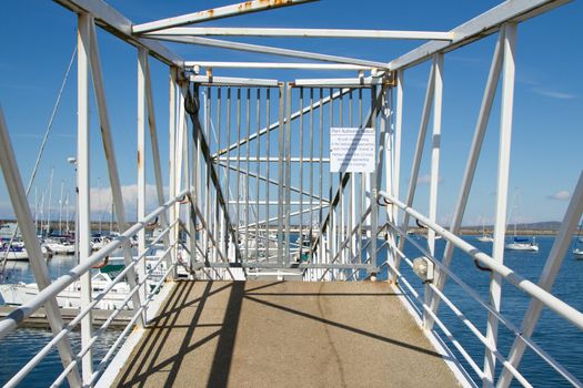 A secure walkway to a marina complex with a raised platform and locked metal gates and bar railings with yachts in the water and a blue sky in the distance.