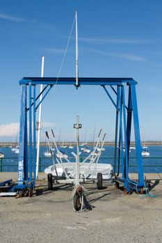 A boat trailer positioned under a blue metal hoist in front of a marina with yachts on a sunny day.