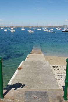 A set of concrete steps leads into the clear waters of a marina with moored yachts and a breakwater in the background.