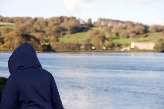 young hoodie wearing teenager exploring by a river in county waterford ireland with large mansion in background