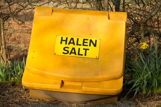 A bright yellow plastic container with a lid and a sign with the words 'Salt Halen' and a daffodil plant next to it.