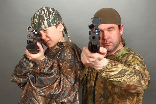 the boy and the adult man in a camouflage with rifles nearby in a shot, aim in a lens