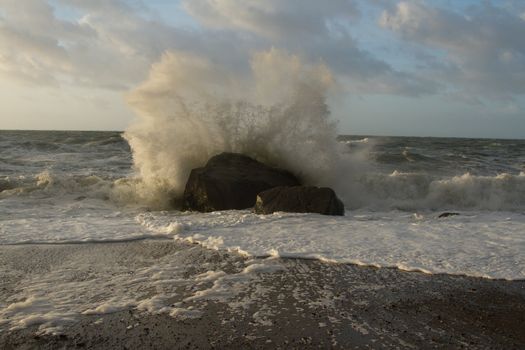 A wave breaks over a boulder on a beach causing the water to create a fan of spray.