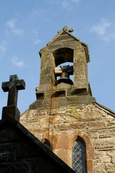 A shaded cross leads to a church bell tower with bell and chain backed by a blue sky.