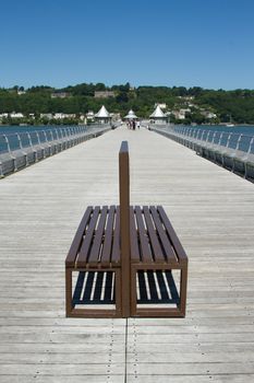 A bouble backed seat made from wood on the decking of a pier which stretching into the distance.