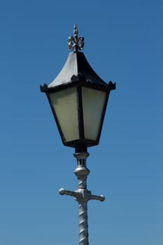 An ornate light with silver post and cross and black metal housing with glass against a blue sky.