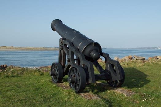 A black painted historic cannon on wheels positioned overlooking a narrow strait of water.