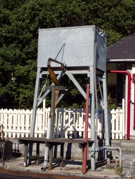 A grey metal steam train water storage tank with pipes and a hose on a raised stand.