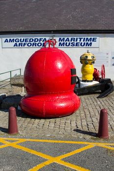 A collection of antique nautical objects on a cobbled area in front of a bilingual sign, Welsh and English, with the words 'Maritime Museum'.