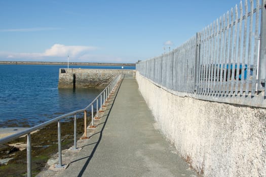 An old quay with steps leading to the sea and a path leading to it with railings and a fence.