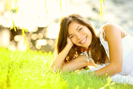 Summer girl in grass smiling happy. Lifestyle image of beautiful young woman in summer dress lying joyful in park on sunny sunshine day. Mixed race Caucasian / Asian Chinese female model.