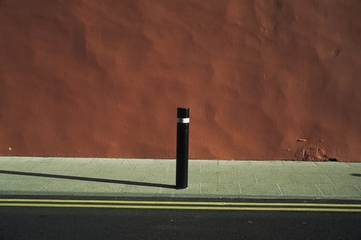 An urban abstract with road, pavement, yellow lines, orange dappled wall and a post with a shadow.