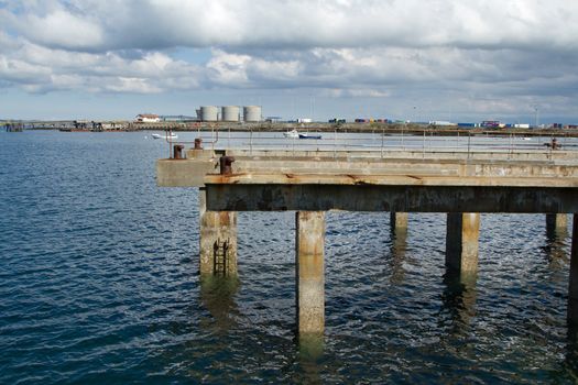 An old dock made of concrete with railings on supports in the sea with storage tanks in the distance.