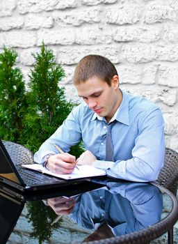 Businessman with laptop in cafe on the street. Makes notes with pen