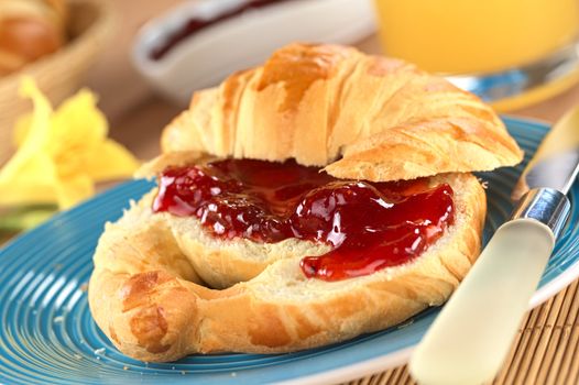 Fresh croissant with butter and strawberry jam on blue plate with orange juice in the back (Selective Focus, Focus on the big strawberry piece in the front)