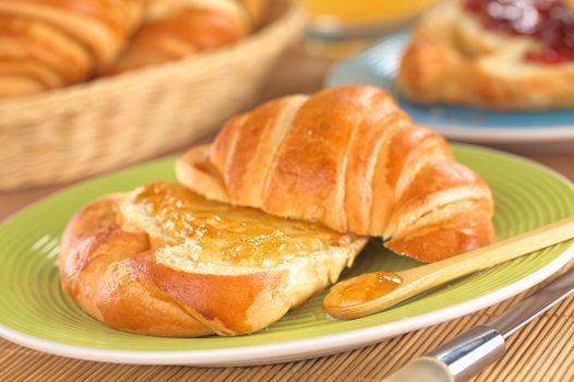 Fresh croissant with butter and orange jam on green plate with a bread basket in the back (Selective Focus, Focus on the front of the orange jam on the croissant and on the wooden spoon)