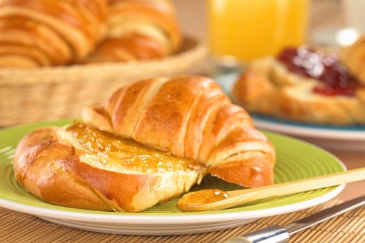 Fresh croissant with butter and orange jam on green plate with juice and bread basket in the back (Selective Focus, Focus on the front of the orange jam on the croissant and on the wooden spoon)