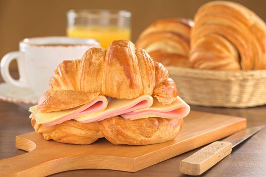 Fresh croissant with ham and cheese on wooden board with coffee, orange juice and bread basket in the back (Selective Focus, Focus on the front of the croissant and the ham and cheese slices)