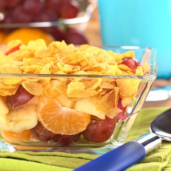 Glass bowl full of fresh fruits (banana, apple, mandarin, grapes) with corn flake cereal on top (Selective Focus, Focus on the mandarin)