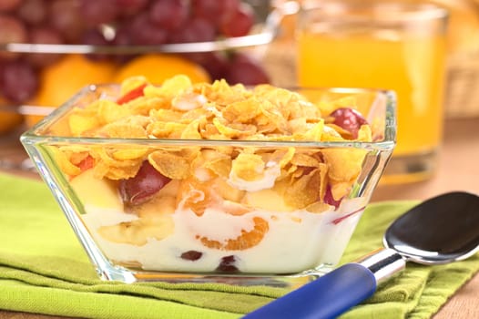 Glass bowl full of fresh fruits (banana, apple, mandarin, grapes) with corn flake cereal and milk (Selective Focus, Focus on the front of the bowl)