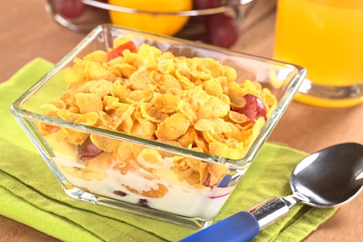 Glass bowl full of fresh fruits (banana, apple, mandarin, grapes) with corn flake cereal and milk with juice in the back (Selective Focus, Focus one third into the corn flakes)