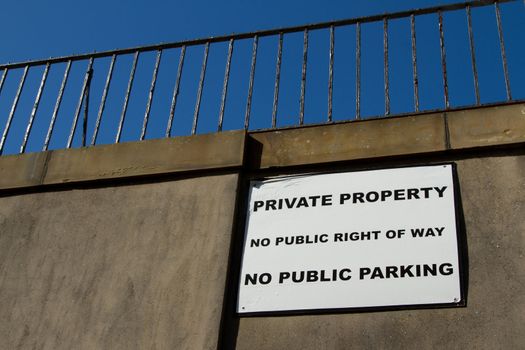 A white square sign with the message 'PRIVATE PROPERTY, NO PUBLIC RIGHT OF WAY and NO PUBLIC PARKING' on a wall with railings against a blue sky.