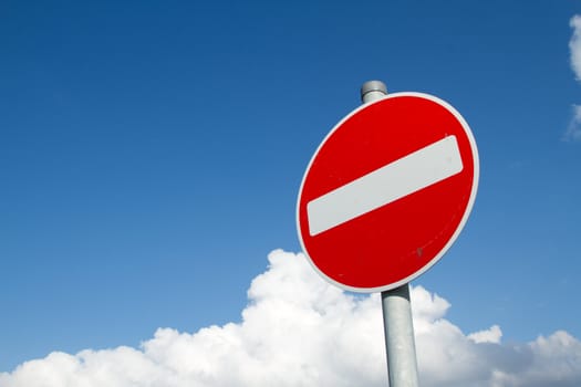 A circular red sign with a white bar indicating 'NO ENTRY' on a grey metal post against a blue cloudy sky.