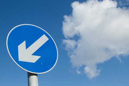 A circular blue sign with a white arrow on a post with a blue cloudy sky in the background.