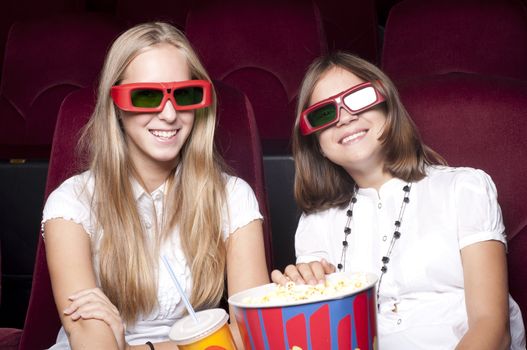 two girls look three-dimensional cinema, sitting in the glasses, eat popcorn, drink drink