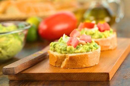 Snack of baguette slices with avocado cream, tomato and onion on wooden cutting board (Selective Focus, Focus on the front of the avocado cream on the first baguette slice)