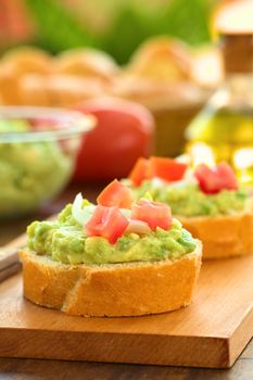 Snack of baguette slices with avocado cream, tomato and onion on wooden cutting board with ingredients in the back (Selective Focus, Focus on the front of the avocado cream on the first baguette slice)