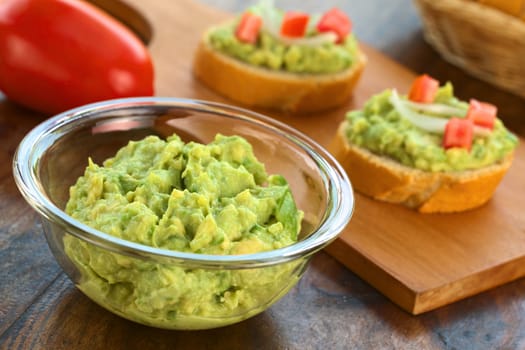 Fresh avocado cream in glass bowl with snacks of baguette with avocado, tomato and onion in the back (Selective Focus, Focus one third into the avocado cream)