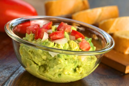 Fresh avocado cream with tomato and onion on top in glass bowl with baguette slices in the back (Selective Focus, Focus on the tomato pieces in the front)