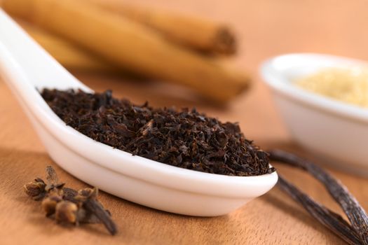 Dried loose black tea on ceramic spoon surrounded by spices such as cloves, vanilla and cinnamon with brown sugar in the back photographed on wood (Selective Focus, Focus one third into the leaves on the spoon)