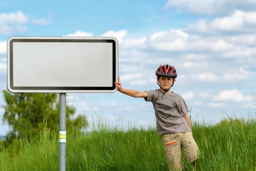 Boy cyclist leaning on blank sign with blue sky and green grass
