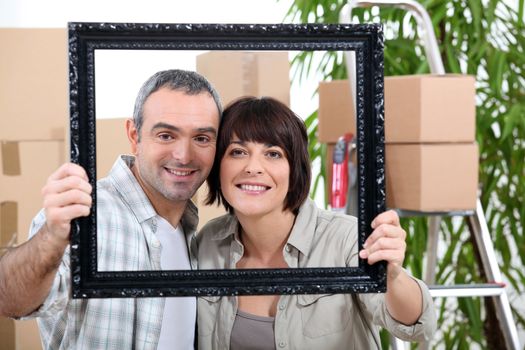 a couple is holding and posing behind a painting frame inside an apartment full of unpacked packages