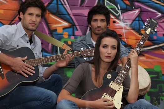 two young men and one young woman playing guitar and djembe in front of a tagged wall