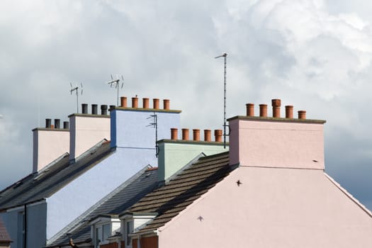 A terraced row of pastel coloured house roofs with chimney pots against a cloudy sky.