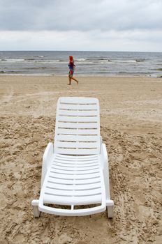 empty plastic white relax chair on beach and young woman walk on sea coastline on cloudy day.