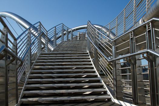 A set of stainless steel steps leads to a bridge with safety hand railings with a blue sky in the background.