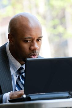 A business man in his early 30s working on his laptop or netbook computer outdoors with a surprised or shocked expression on his face.