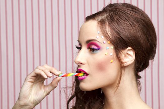 Young woman with colorful makeup and star candy glued to her face, bitting a candy cane